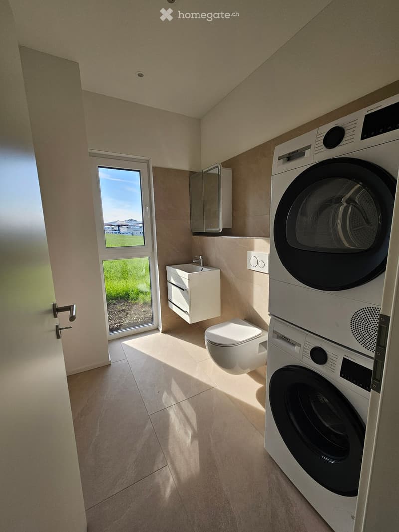 Modern bathroom with a toilet, sink, mirror, and a stacked washing machine/dryer unit. Natural light streams in from a large window.