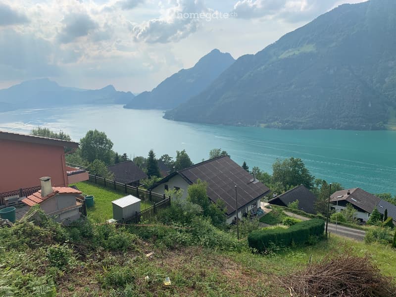 A scenic view of a lakeside area with several houses, surrounded by lush greenery and mountains in the background.