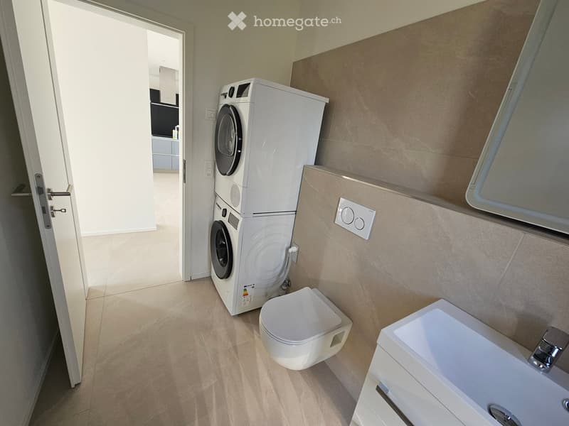 Modern bathroom with a toilet, sink, and stacked washing machine. Light-colored tiles and a clean, minimalist design.