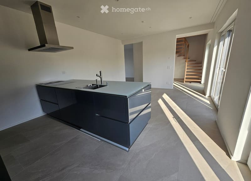 Modern kitchen with dark grey cabinets, a central island with a sink and faucet, and a range hood above. The floor is tiled, and there is a staircase visible in the background.