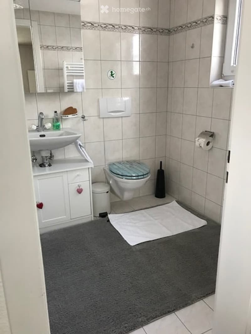 A small bathroom with beige tiled walls and floor, a white sink with a mirror above it, a toilet with a blue seat cover, a white cabinet under the sink, a gray bath mat, and a towel hanging on the wall.