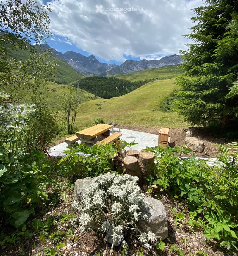 A serene outdoor garden area with a wooden picnic table, benches, log stumps, and lush greenery, overlooking a picturesque mountain landscape.
