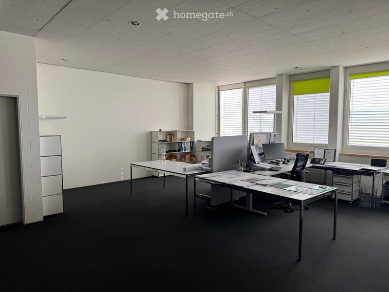 Modern office space with desks, chairs, computers, and shelves. Large windows with blinds provide natural light.