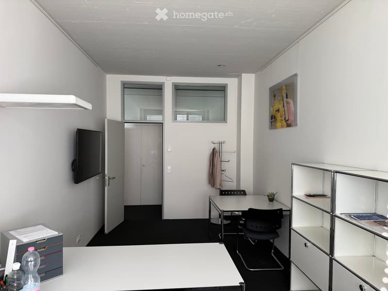 Modern office space with white walls, black flooring, and a large window. Includes a desk with chairs, a screen, a shelf, and a small plant.