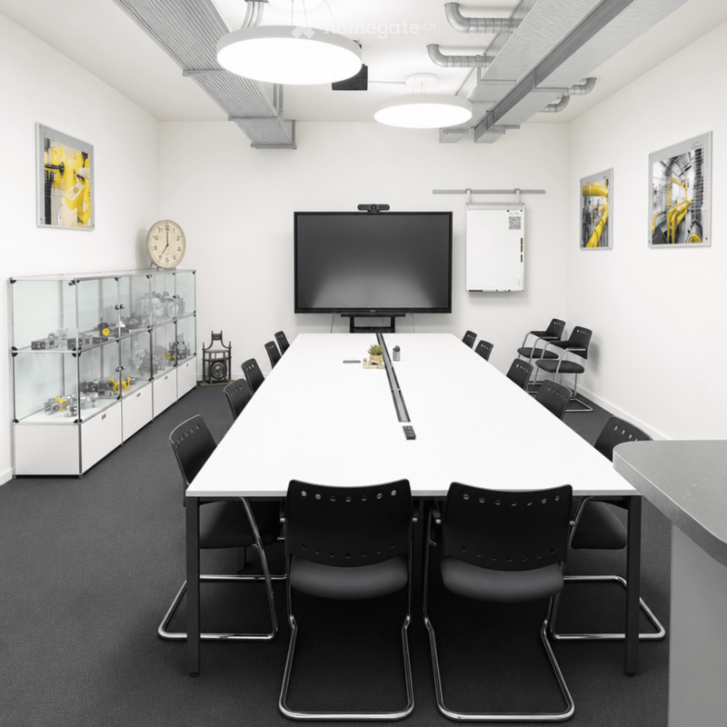 Modern conference room with a long white table, black chairs, a large screen, a clock, and a glass cabinet with mechanical parts.