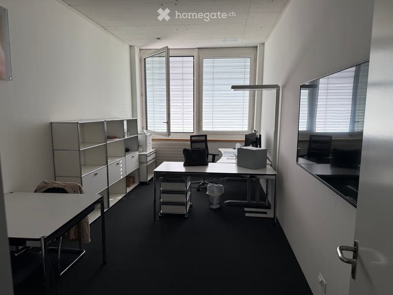Modern office space with two desks, office chairs, a laptop, printer, and a cabinet. Large windows with blinds provide natural light.