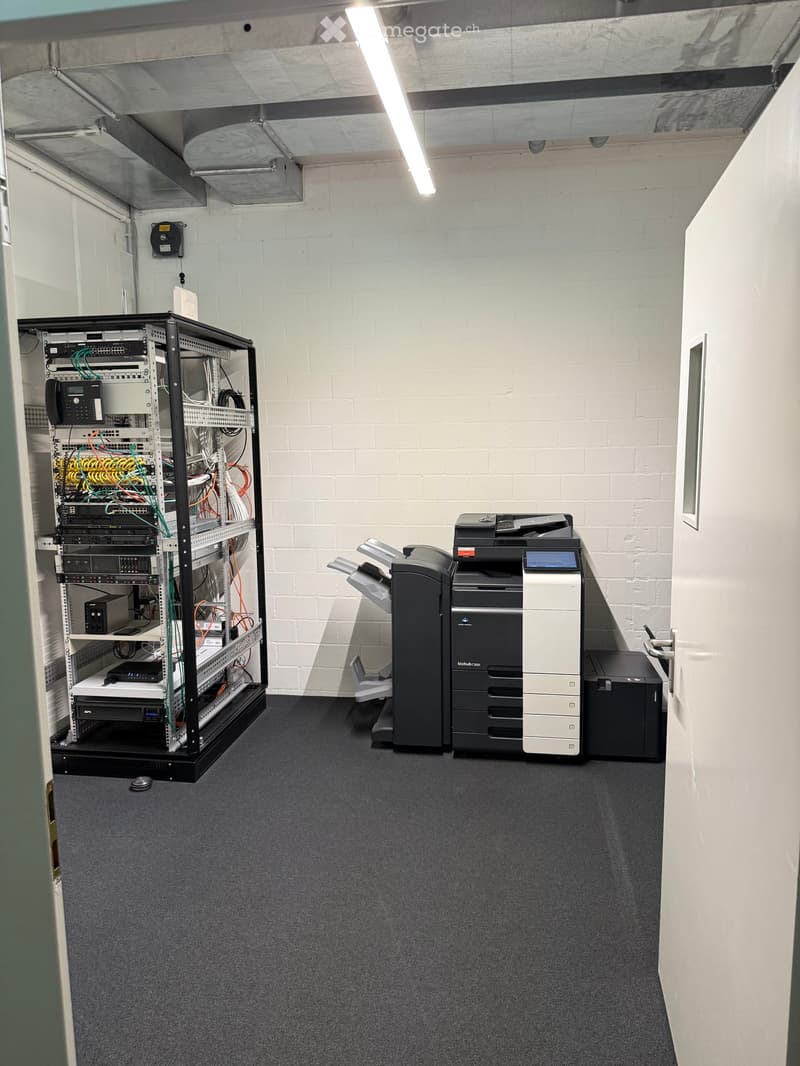 A small, organized room with a server rack filled with networking equipment and a large printer on the floor. The walls are white brick, and the floor is carpeted.