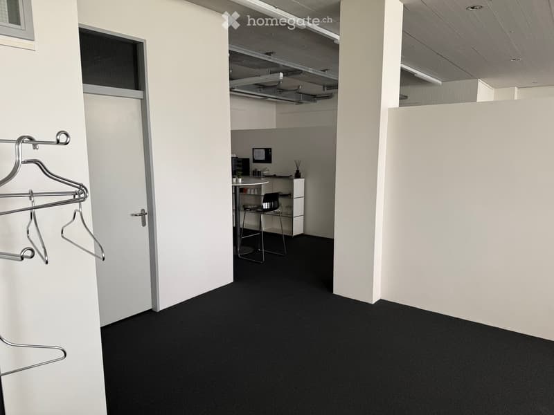 A modern, minimalistic room with white walls, black carpet, and a small kitchenette area featuring a table, chair, and cabinet.