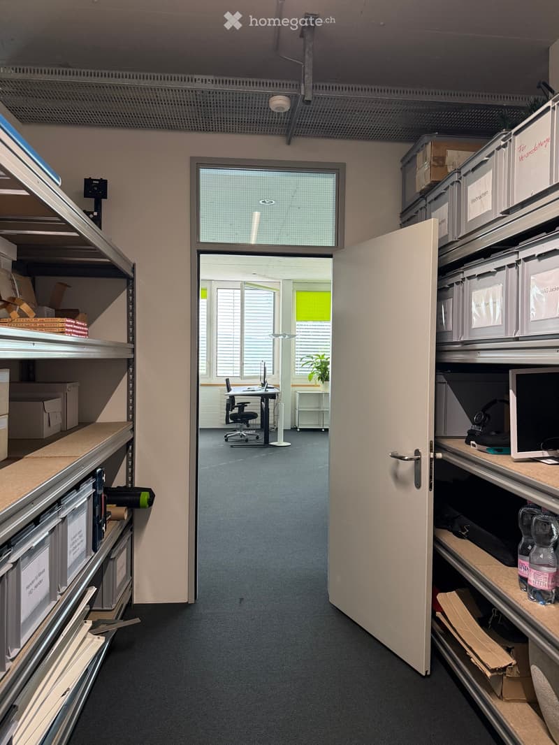 Office space with a desk, chair, computer screen, shelves filled with storage bins, and a water bottle on a shelf.
