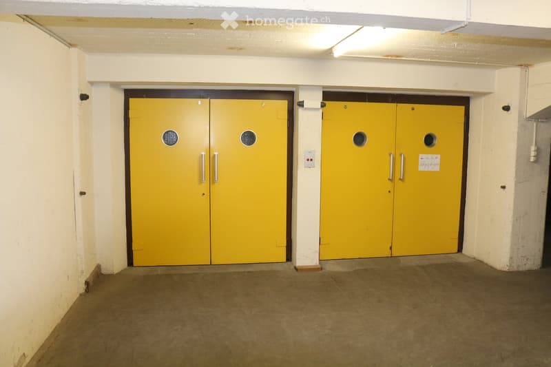 Empty underground garage area with two large yellow garage doors, concrete floor, and white walls.