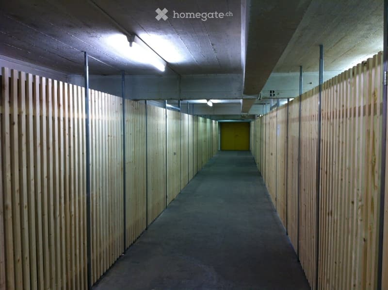 A long, narrow hallway with wooden panel walls, concrete floor, and fluorescent lighting. The corridor leads to a yellow door at the far end.