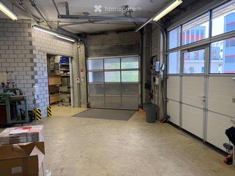 Empty garage with large industrial doors, concrete floor, exposed brick wall, and storage area with shelves and tools.