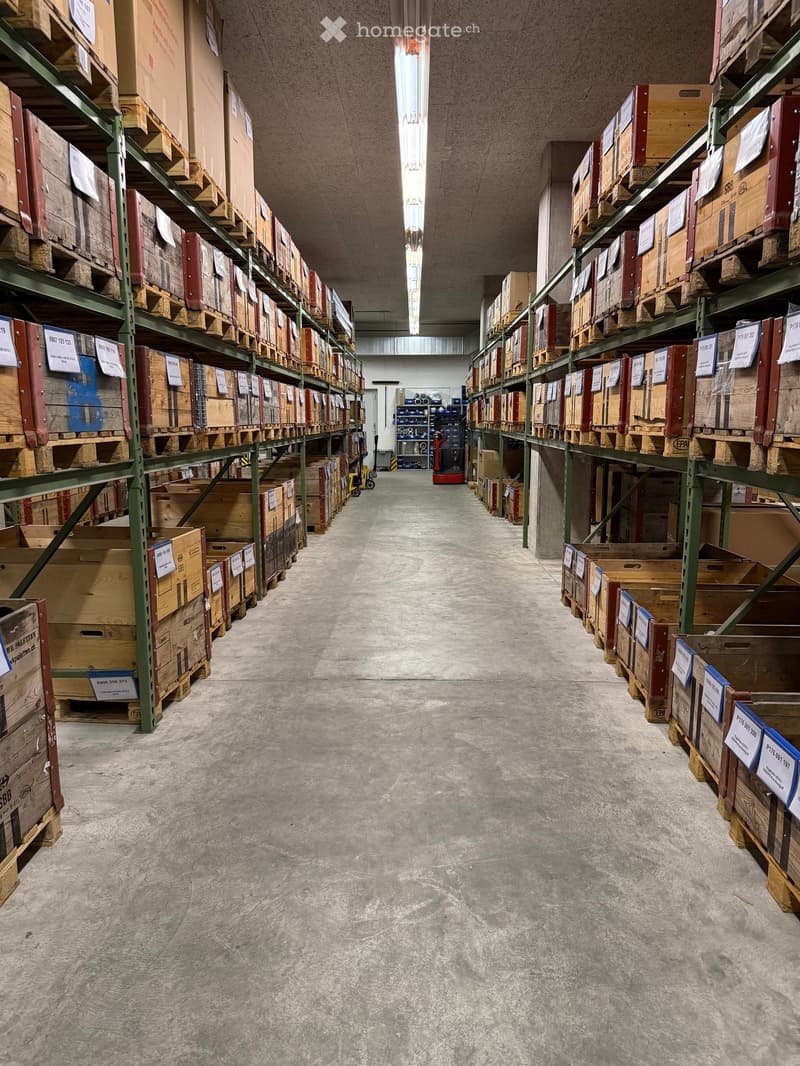 A long, wide corridor with high ceilings and industrial lighting, lined with green metal shelving units holding numerous wooden crates and boxes.