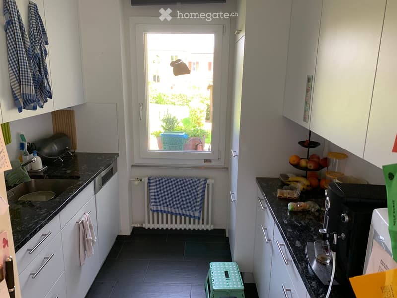 Modern kitchen with white cabinets, dark granite countertops, and stainless steel appliances. Features include a sink, microwave, and a small green stool near the window.