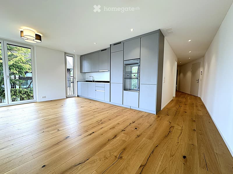 Unfurnished Room. Empty room with light grey cabinets, a sink, and a built-in oven. The floor is made of light wooden planks, and the walls are white. There are large windows with a view of greenery outside.