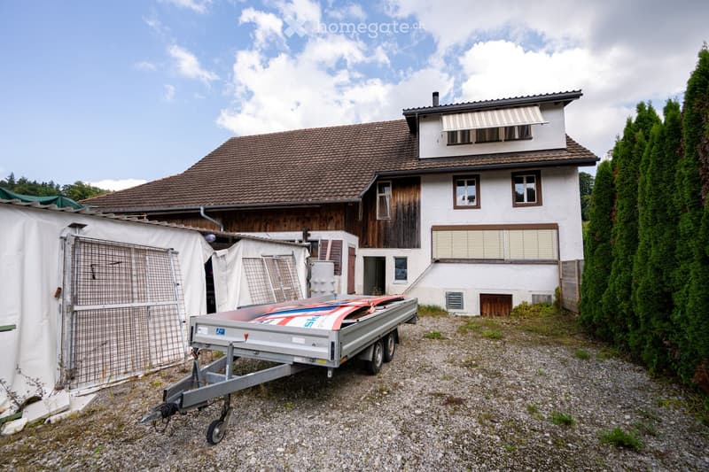 Two-story house with a brown roof and white walls, a trailer parked in front, an attached garage, two white canopies on the left, and a window with shutters on the right.