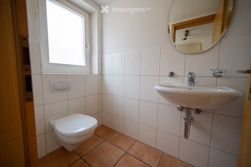 A small bathroom with white walls and brown tiled floor. There is a toilet, sink, and a mirror mounted on the wall.