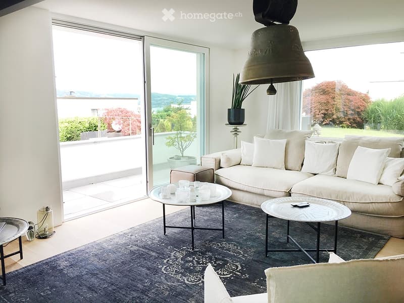 Bright living room with large windows and a balcony view, featuring a cream-colored sectional sofa, coffee table, side table, and a patterned rug.
