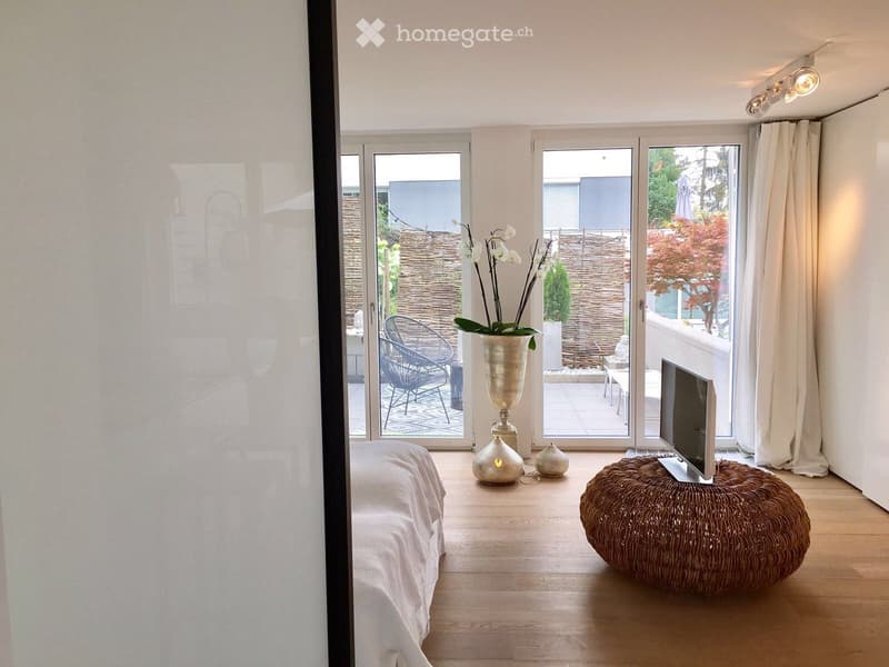 Modern bedroom with wooden flooring, large windows, a bed with white linens, a wicker stool with a TV, a vase with flowers, and a chair visible through the open sliding doors to a balcony.