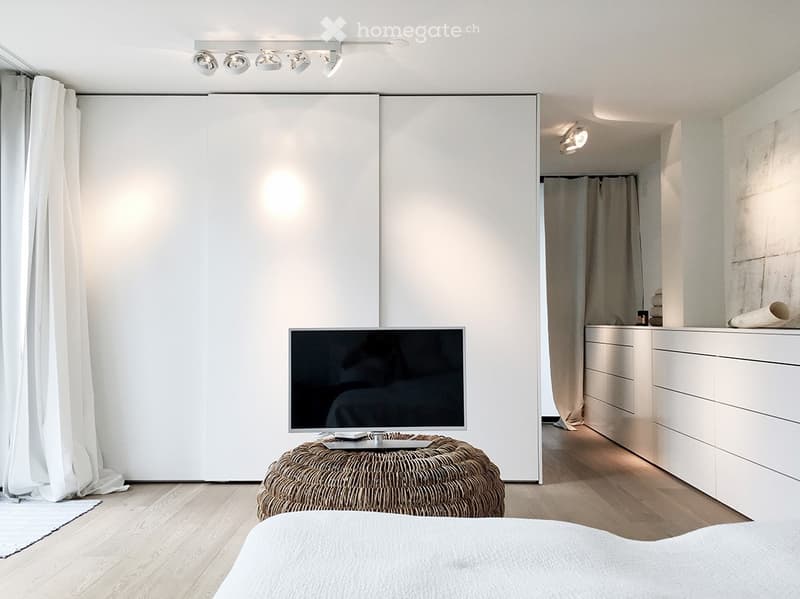 Modern, minimalist bedroom with a large bed, wicker stool, flat-screen TV, and a walk-in closet with a wardrobe and mirror.