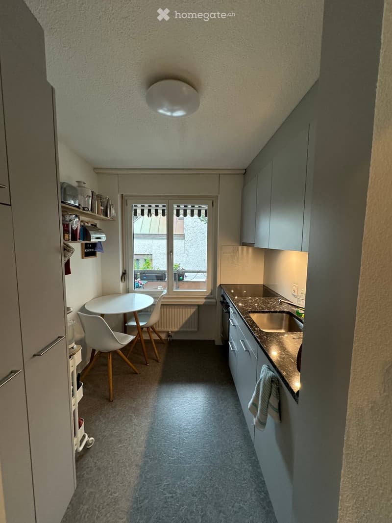 Compact kitchen with a round white table, two white chairs, a sink, and built-in cabinets.