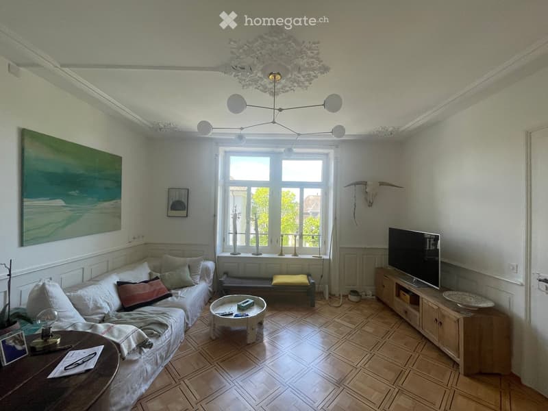 Bright living room with wooden flooring, large window, modern chandelier, sofa, coffee table, TV, and decorative items.