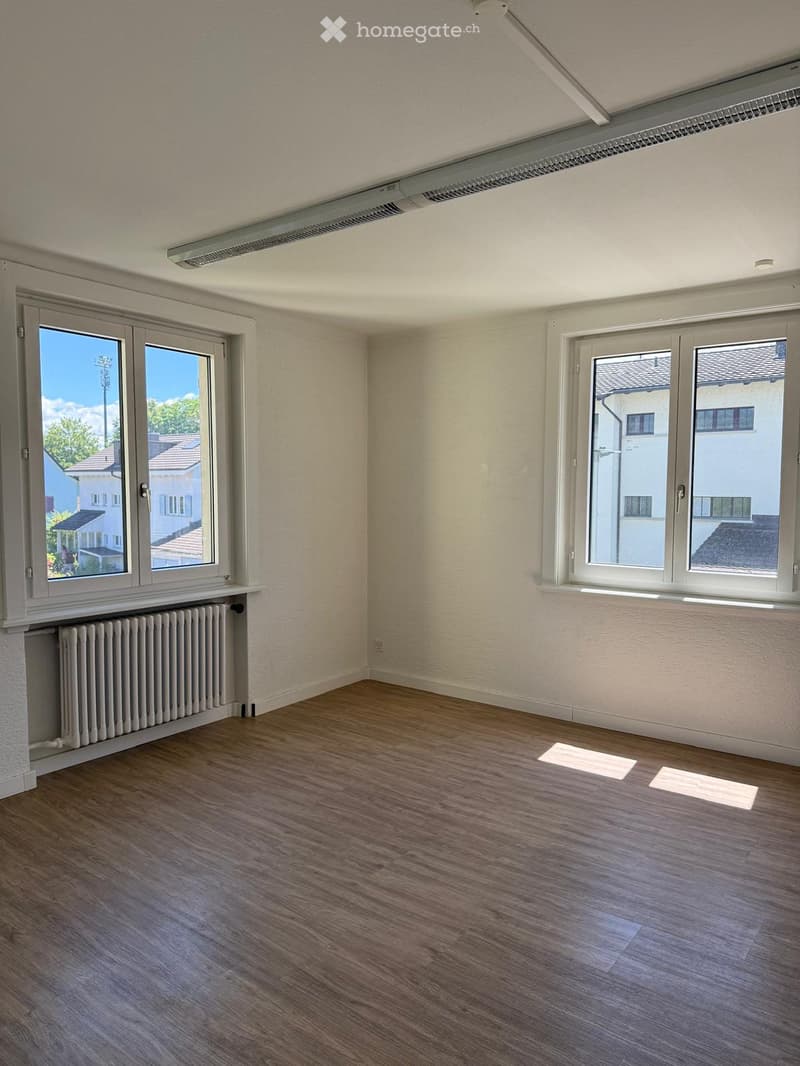 Empty room with wooden flooring, white walls, large windows, radiator, and visible electrical outlets.