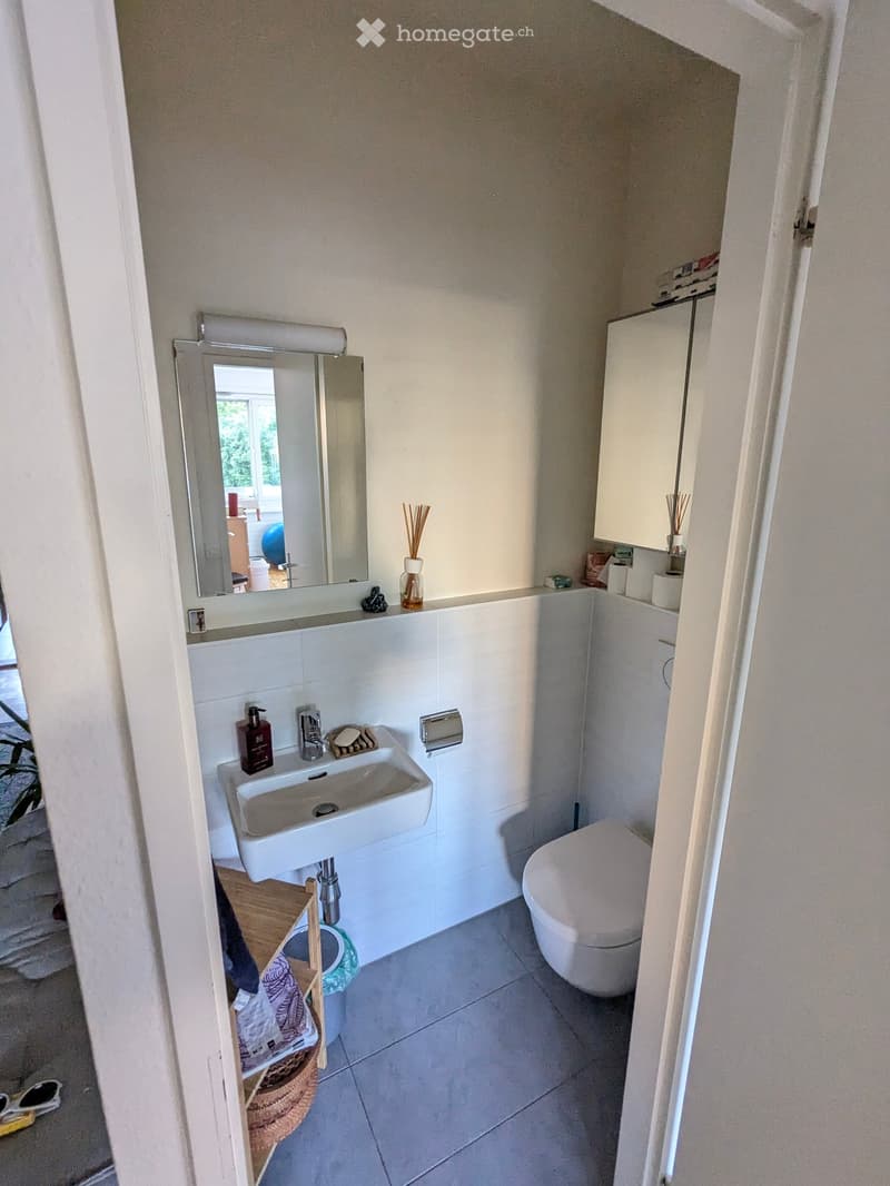 Modern bathroom with white tiles, a sink, toilet, mirror, and cabinet. Includes a small wooden shelf with a basket and a few items.