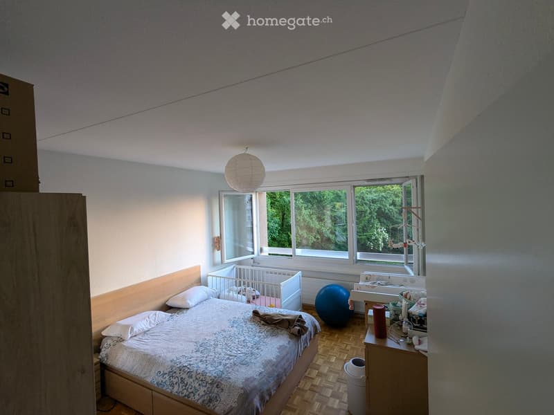 A well-lit bedroom with a wooden bed, nightstand, and a baby crib. The room features large windows with a view of greenery, a wooden floor, and a small table with various items.