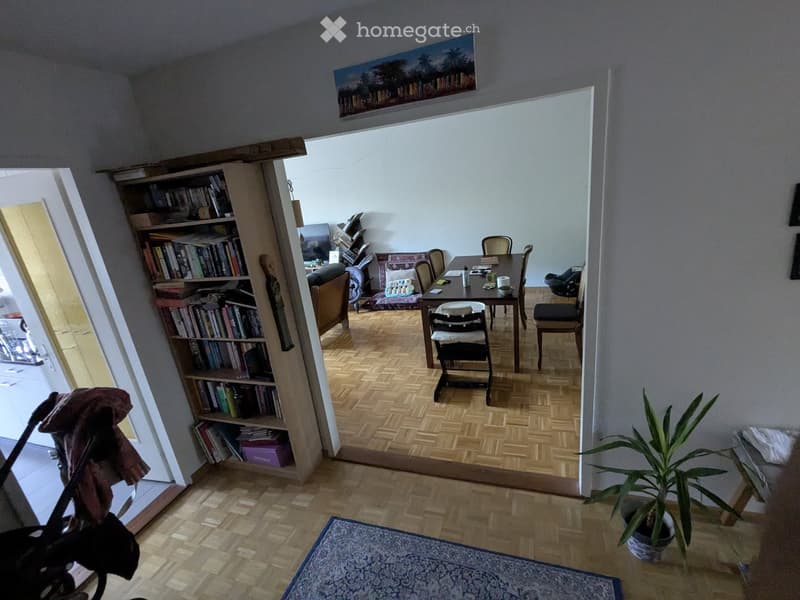 A furnished room with a wooden bookshelf, a dining table with chairs, a sofa, and a potted plant. The room features wooden flooring and a patterned rug.