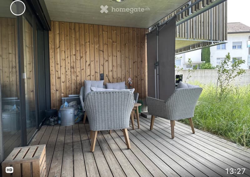 Wooden balcony with wicker chairs, small table, and wooden decking. Features a wooden wall and a retractable awning.