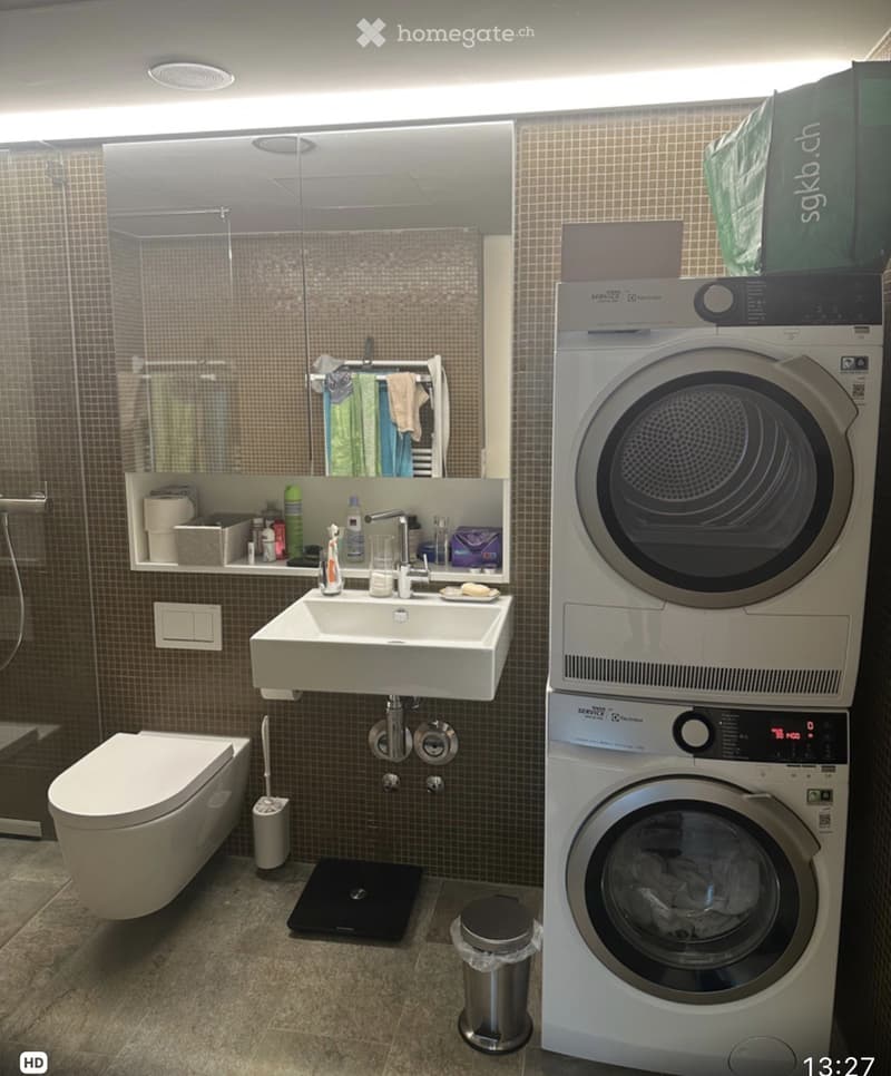 Modern bathroom with a white sink, toilet, washing machine, dryer, and a glass-enclosed shower area. The walls are tiled, and there is a towel rack with towels.