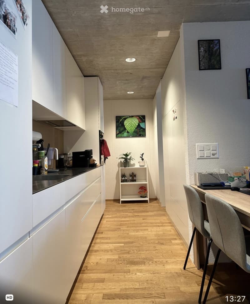 Modern kitchen area with white cabinets, a coffee machine, a small shelf with plants and decorative items, and a dining area with a table and chairs.