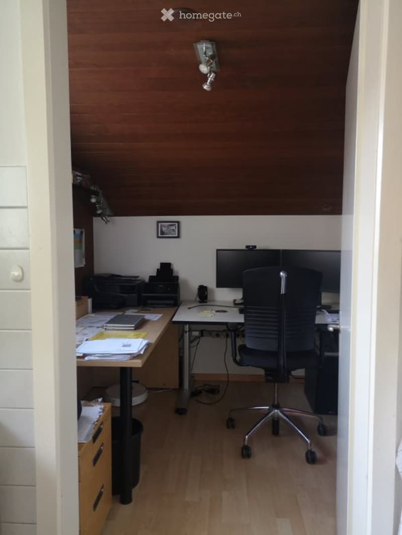 A small, modern office space with a wooden slanted ceiling, a desk with a laptop, printer, and papers, a black office chair, and a wall-mounted lamp.