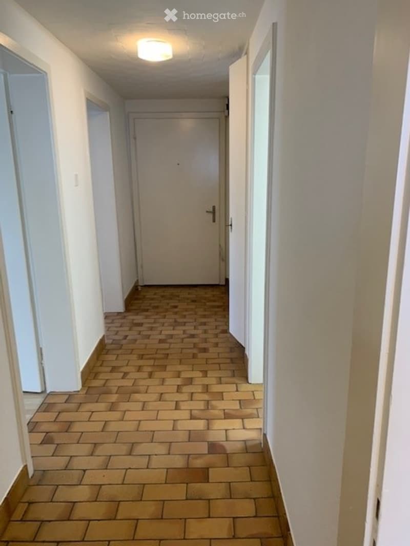 A narrow hallway with a tiled floor and white walls, featuring a closed door at the end and several open doorways along the sides.