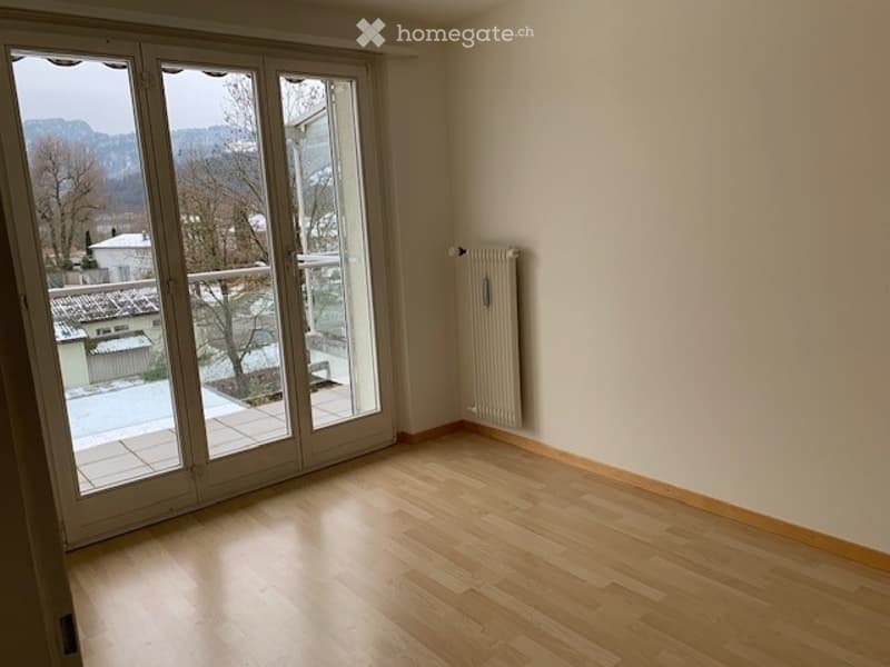 Empty room with wooden flooring, large glass sliding doors leading to a balcony, and a radiator on the wall.