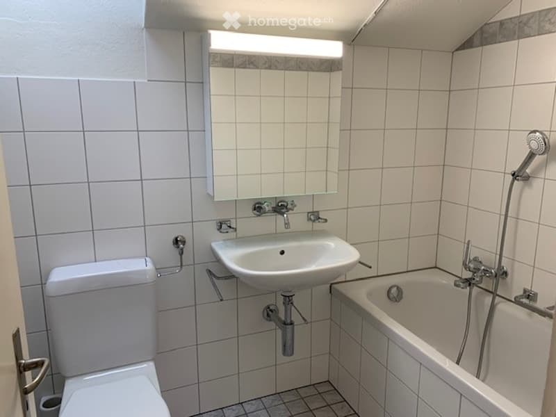Modern bathroom with white tiled walls and floor, featuring a toilet, sink, bathtub, and a mirror above the sink.