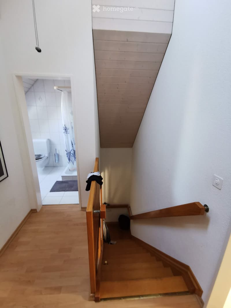 A narrow wooden staircase with a wooden railing leads up to an attic space. A small bathroom is visible at the end of the hallway, featuring white tiles and a toilet.