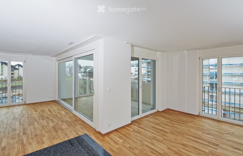 Empty room with wooden flooring, white walls, large glass sliding doors leading to a balcony, and a small rug on the floor.