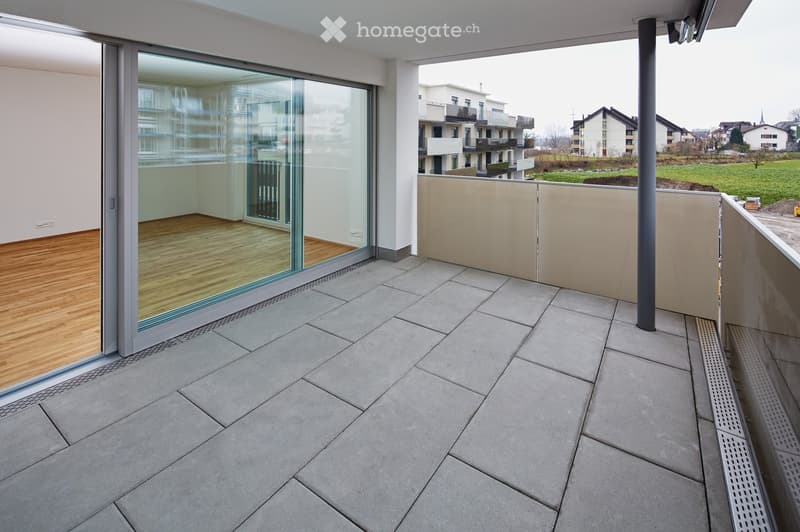Balcony with grey tiled floor, glass sliding door leading to a wooden-floored room, beige railing, and a view of surrounding buildings and green fields.