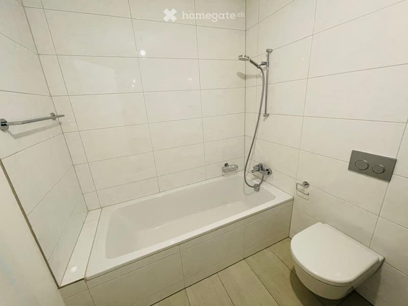 Modern bathroom with white tiled walls and floor, featuring a bathtub, toilet, showerhead, and towel rack.