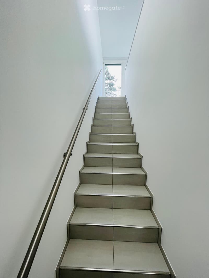 Modern staircase with grey tiled steps and a stainless steel handrail, leading to a bright window at the top.
