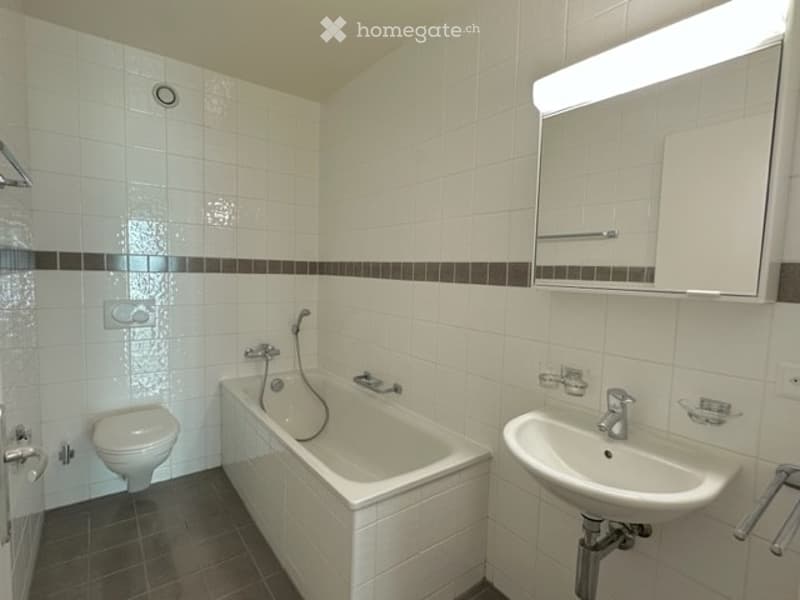 Modern bathroom with white tiled walls, a bathtub, toilet, sink, and a large mirror with built-in lighting.