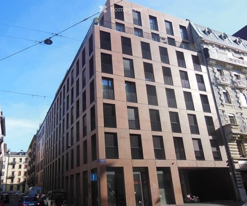Modern multi-story building with a brown facade, numerous windows, and balconies. The building is situated in an urban area with other structures visible in the background.