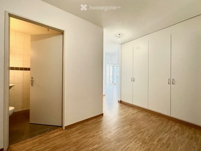 Empty room with light wooden flooring, white walls, built-in white cabinets, and an open doorway leading to a tiled bathroom.