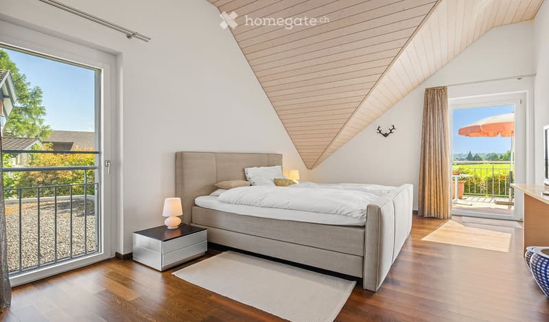 Modern bedroom with wooden floors, large windows, a bed with pillows and a headboard, nightstand with lamp, and a view of the garden.