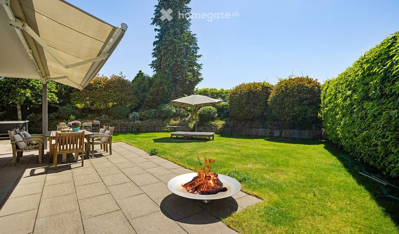 Well-maintained garden with a fire pit, lounge chairs, dining table, and umbrella, surrounded by greenery and a paved patio.