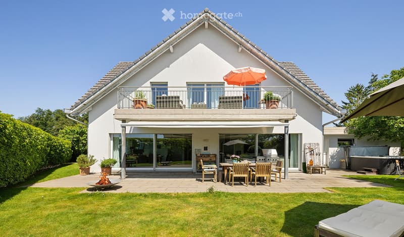 Modern two-story house with a spacious garden, patio seating area, outdoor dining table, and a balcony with an umbrella.