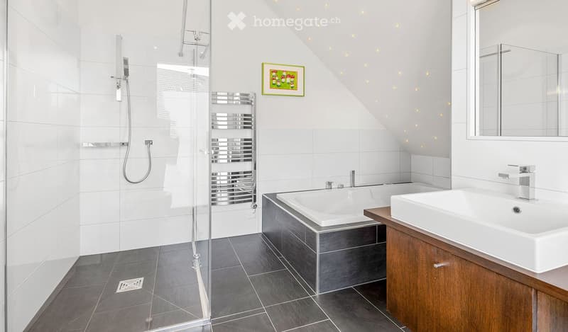 Modern bathroom with a glass-enclosed shower, bathtub, double sink vanity, mirror, and a radiator. The space features white tiles and a sloped ceiling with recessed lighting.