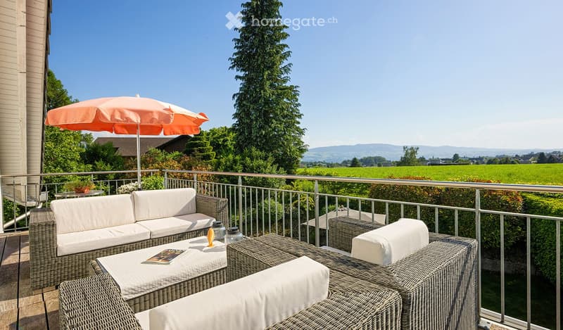 Balcony with wicker furniture, including a sofa, table, chairs, and an umbrella, overlooking a scenic countryside view.